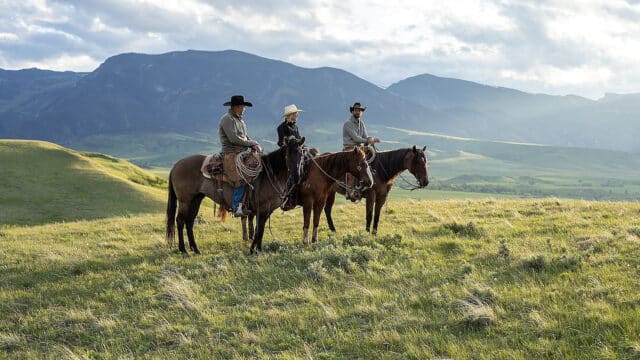 Three horseback riders on lush rolling grasslands at a northern Wyoming ranch, with the Bighorn Mountains in the background.